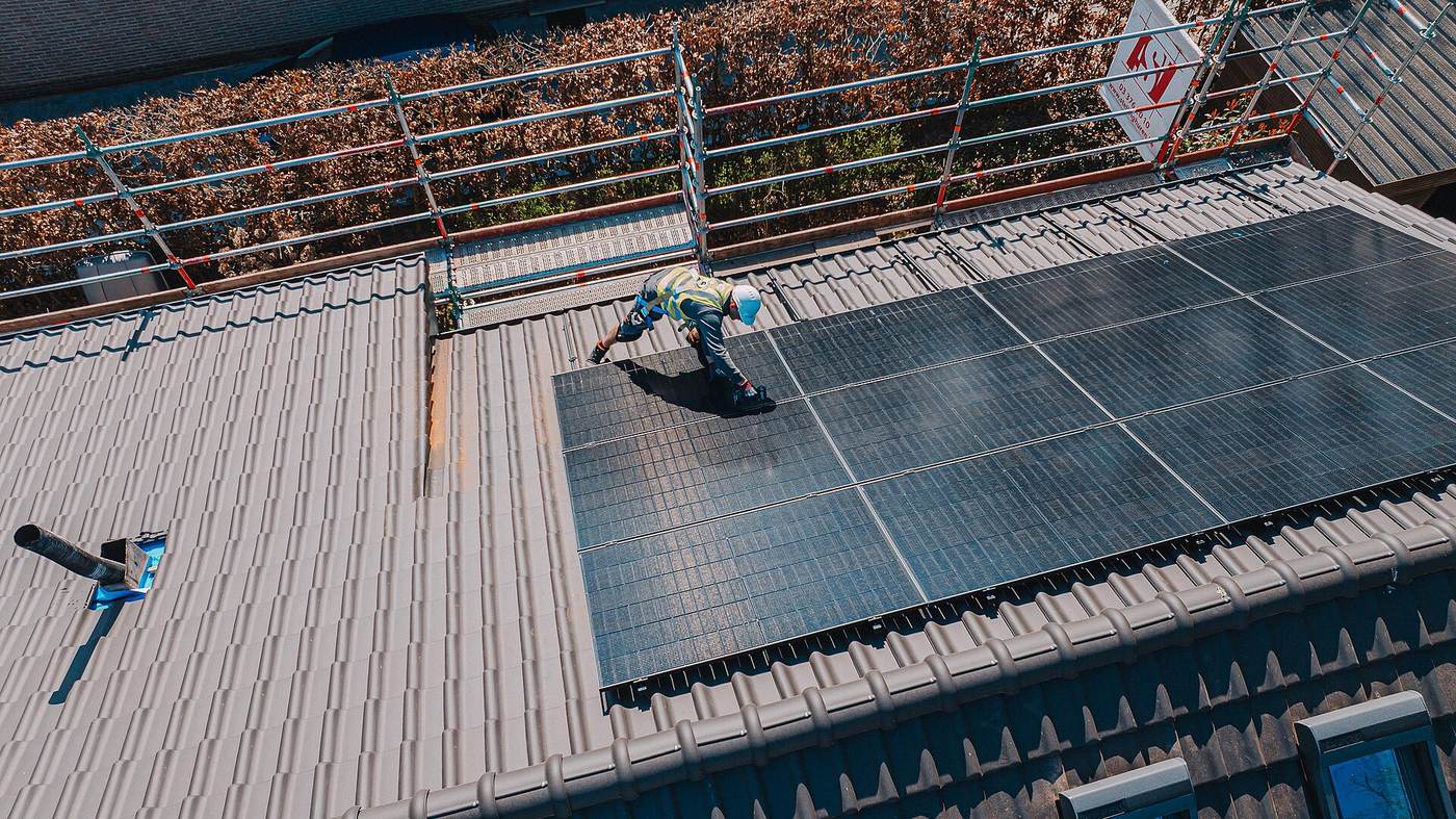 Installateur werkt op een pannendak naast een rij pas geplaatste zonnepanelen, achter een dakrandbeveiliging