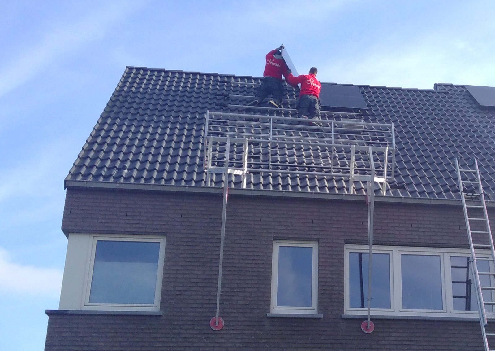 Twee installateurs in rode werkkledij plaatsen zonnepanelen op een donker pannendak, met valbeveiliging en ladder